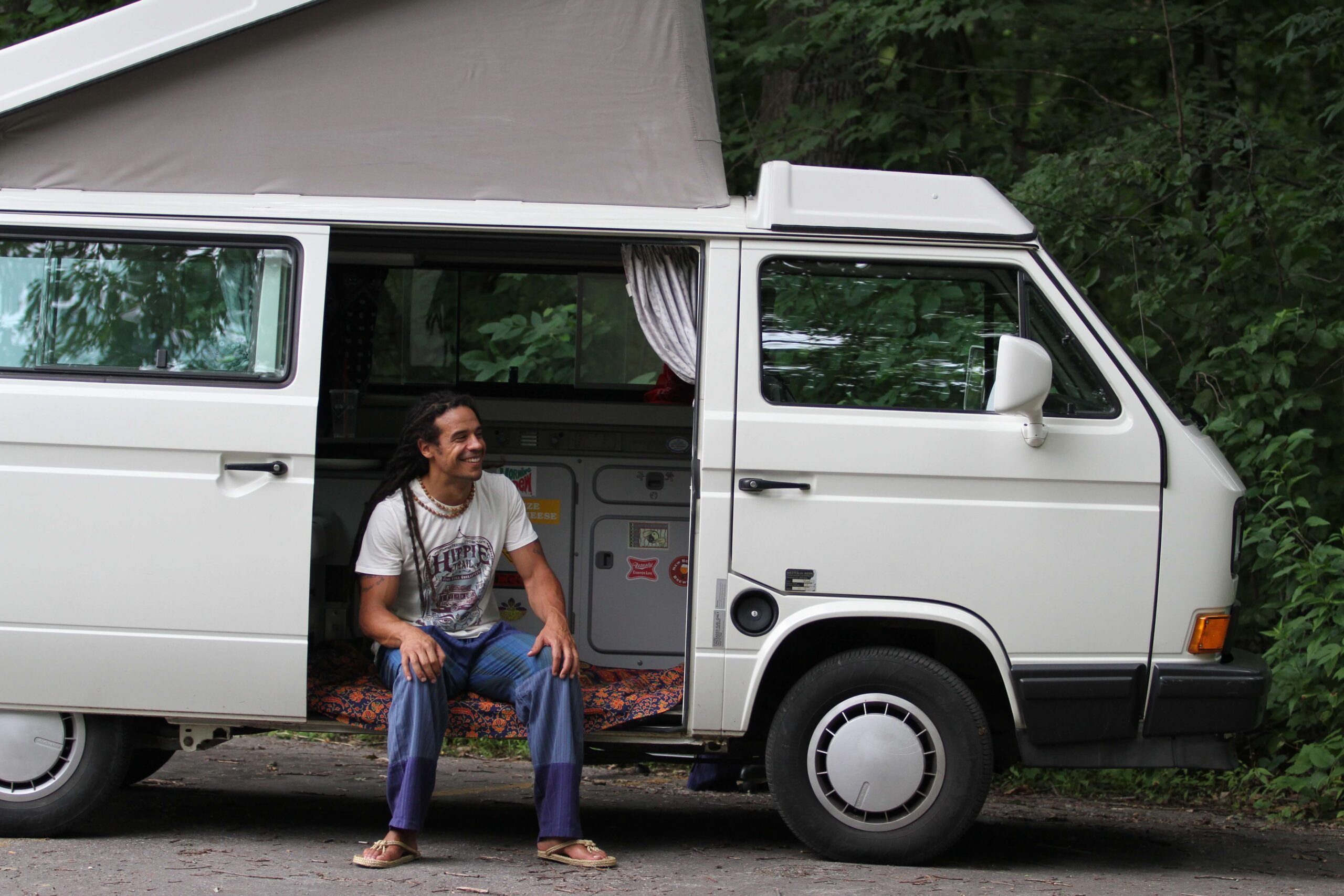 A person sits in the doorway of a white camper van parked on a road, surrounded by trees and greenery. Soul Flower Blog: Hippie Style & Kind Living