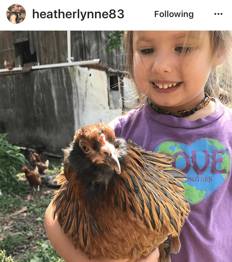 A kind young girl in a purple LOVE shirt smiles while holding a brown chicken outdoors, with more chickens and a rustic building creating a boho atmosphere in the background. Soul Flower Blog: Hippie Style & Kind Living
