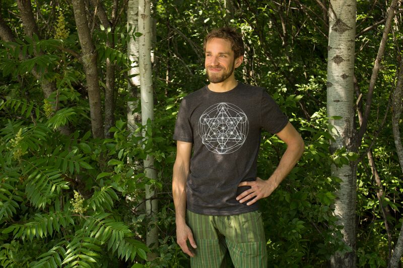 A man stands in a forested area with one hand on his hip, wearing a dark geometric-print t-shirt and green striped pants—ready to kickstart his week with a Monday Motivation Playlist. Soul Flower Blog: Hippie Style & Kind Living