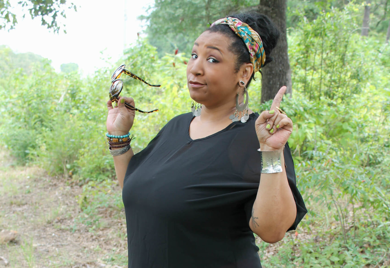 A woman in a funky black top, colorful headband, and jewelry stands outdoors holding sunglasses in one hand and raising her index finger on the other. Trees and eco greenery fill the background. Soul Flower Blog: Hippie Style & Kind Living