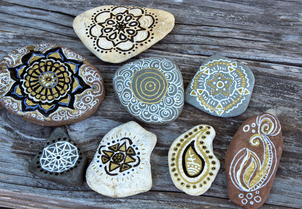 Eight smooth stones with intricate boho mandala and paisley patterns painted in black, white, and gold, arranged on weathered wooden boards for an earthy, artistic touch. Soul Flower Blog: Hippie Style & Kind Living