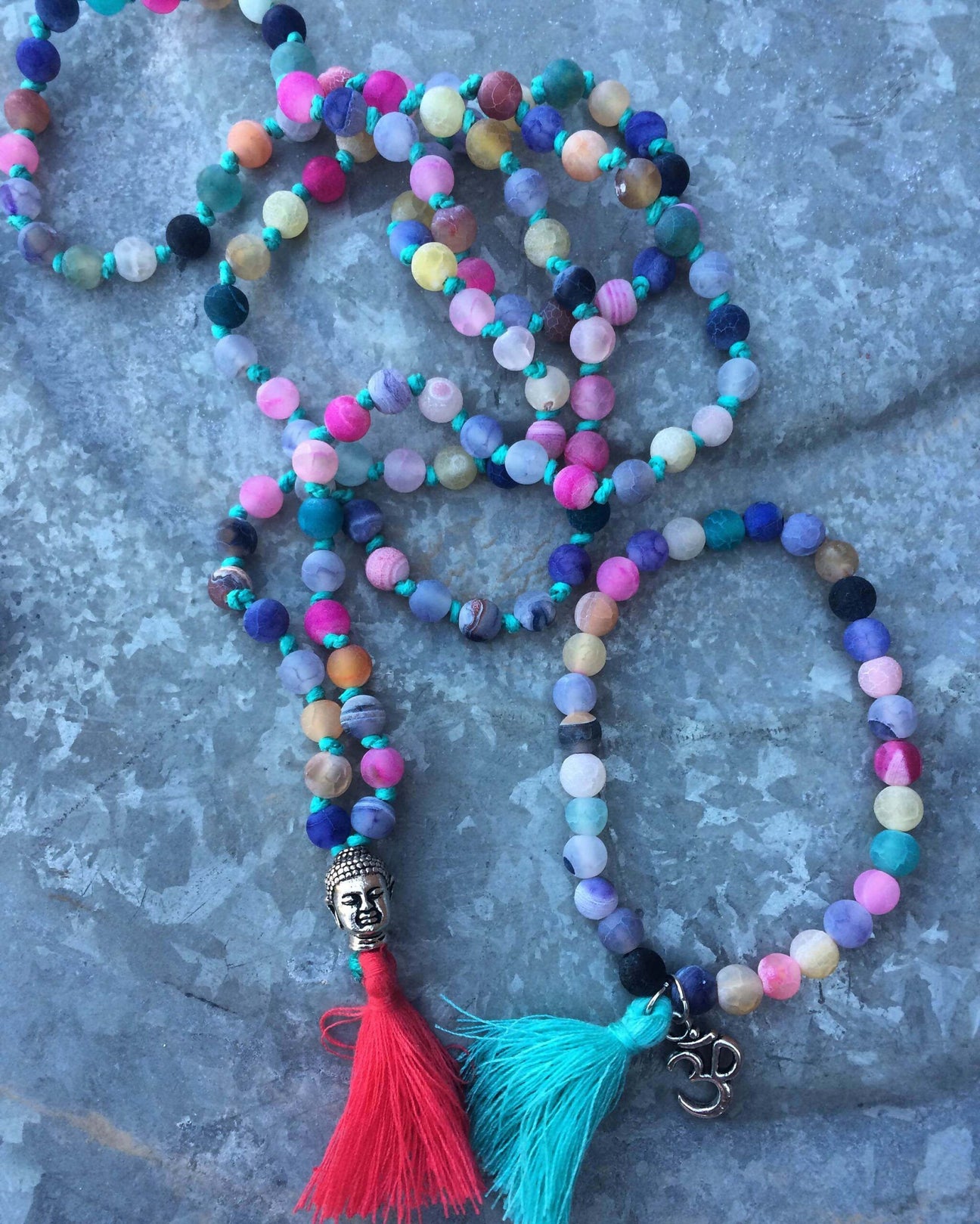 A beaded necklace from Mantra & Malas with multicolored round beads, a silver Buddha charm and red tassel, and another featuring a silver Om charm with blue tassel, elegantly displayed on a stone surface. Soul Flower Blog: Hippie Style & Kind Living