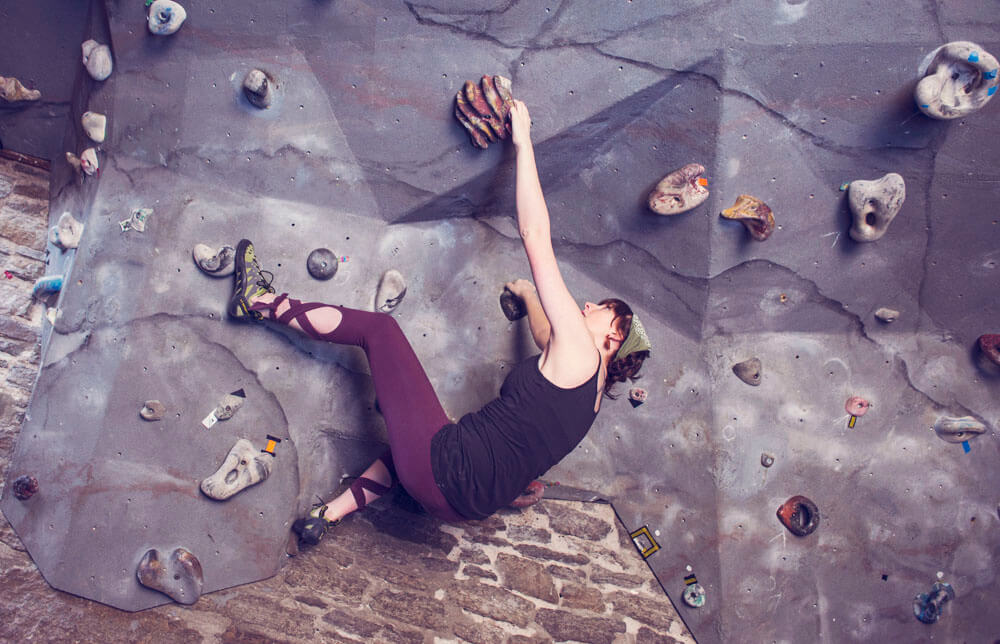 A person in boho athletic clothing climbs an indoor bouldering wall, gripping handholds and wearing climbing shoes against a textured surface. Soul Flower Blog: Hippie Style & Kind Living