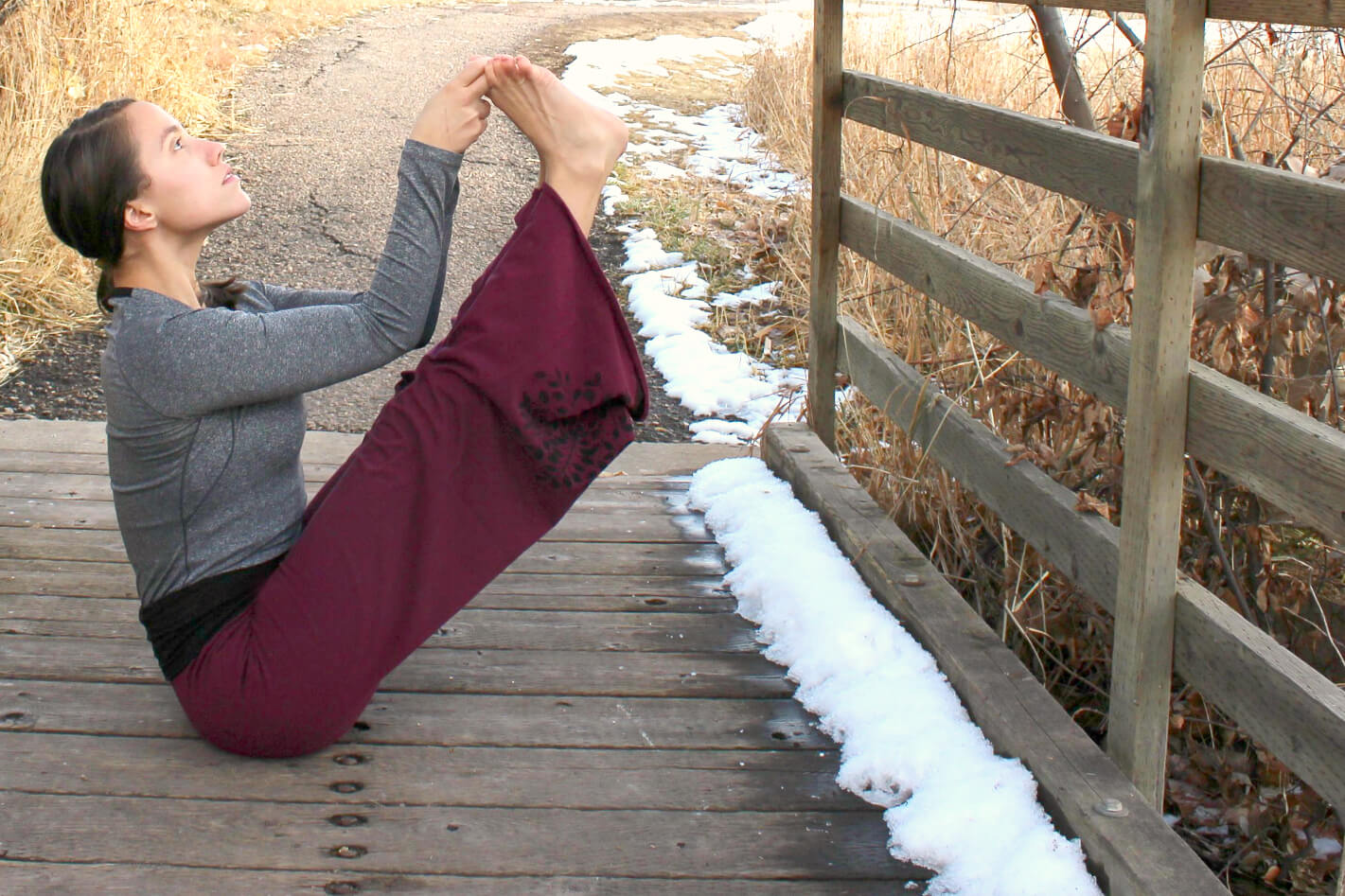 A person in a gray long-sleeve shirt and maroon pants practices a seated yoga pose on a wooden bridge, embracing an eco-boho vibe amid patches of snow and dry grass. Soul Flower Blog: Hippie Style & Kind Living