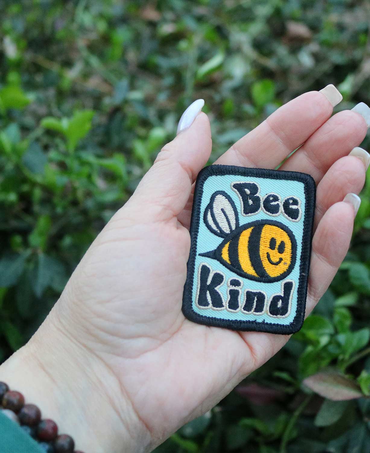 A Soul Flower Bee Kind Patch held in a hand in front of greenery outdoors.