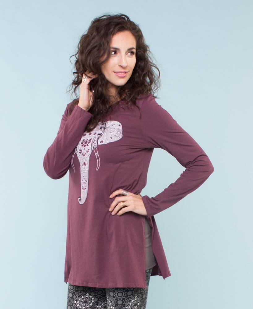 Woman with wavy brown hair wearing a long-sleeve, maroon tunic featuring a kind elephant graphic, standing against a light blue background. Soul Flower Blog: Hippie Style & Kind Living