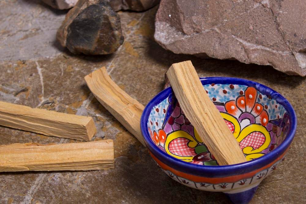 Three palo santo wood sticks rest on a stone surface, one placed inside a colorful ceramic bowl. With rocks in the background, this eco-inspired scene brings a touch of boho charm to any space. Soul Flower Blog: Hippie Style & Kind Living