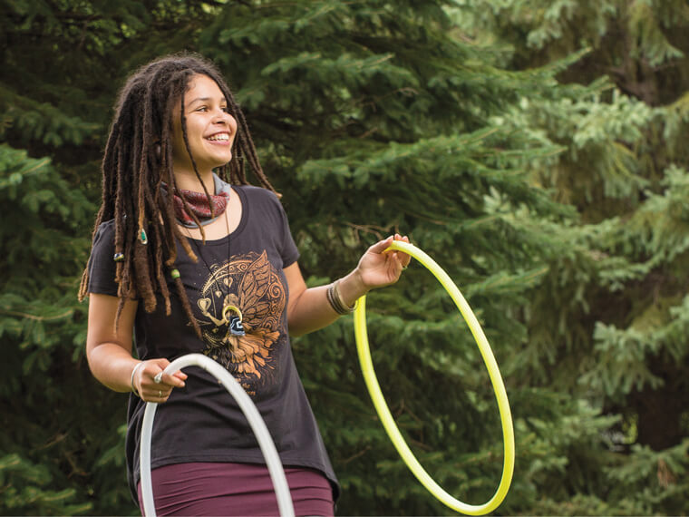 A funky person with long dreadlocks smiles while holding two hula hoops, one white and one yellow, standing outdoors in front of evergreen trees. Soul Flower Blog: Hippie Style & Kind Living