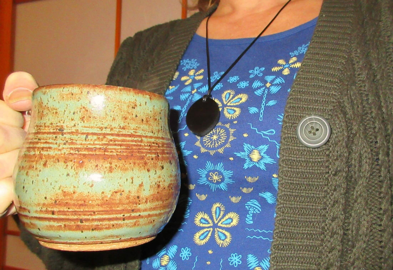 Person wearing a blue floral shirt and dark cardigan holds a ceramic mug with brown and green hues in their left hand. A round black pendant hangs on a necklace, adding a subtle boho touch to the cozy scene. Soul Flower Blog: Hippie Style & Kind Living