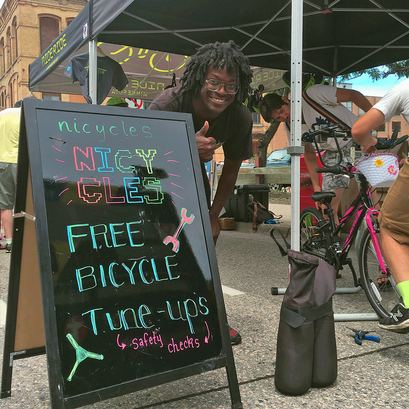 A person in a boho hippie t-shirt gives a thumbs up behind a sign reading Nicycles Free Bicycle Tune-ups (safety checks) at an outdoor event with bikes and tents, creating an eco-friendly vibe. Soul Flower Blog: Hippie Style & Kind Living