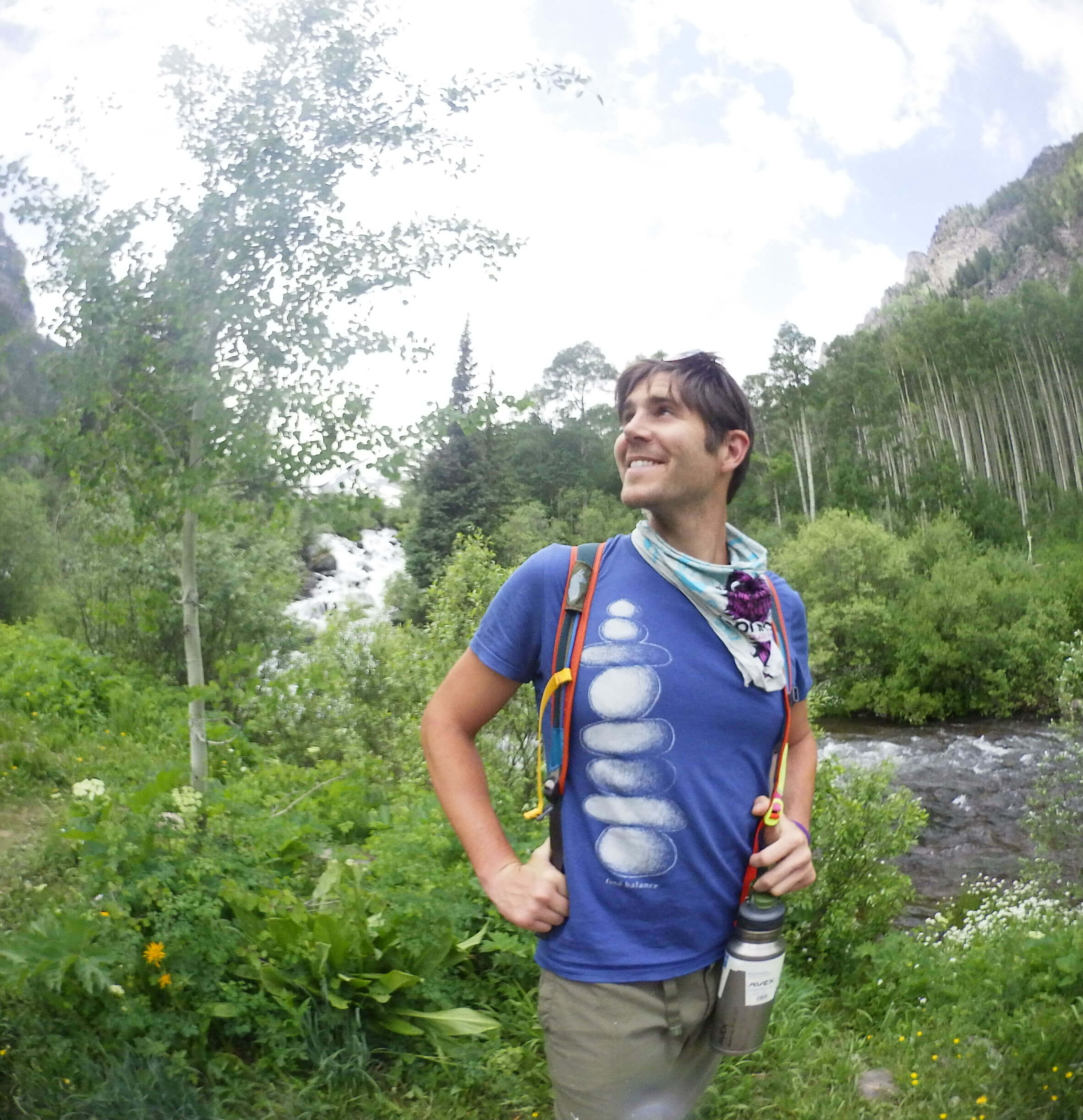 A person wearing a blue t-shirt and backpack stands smiling by a lush green riverside, perhaps reflecting on Understanding (Capital F) Music Festivals, with trees and a waterfall in the background. Soul Flower Blog: Hippie Style & Kind Living