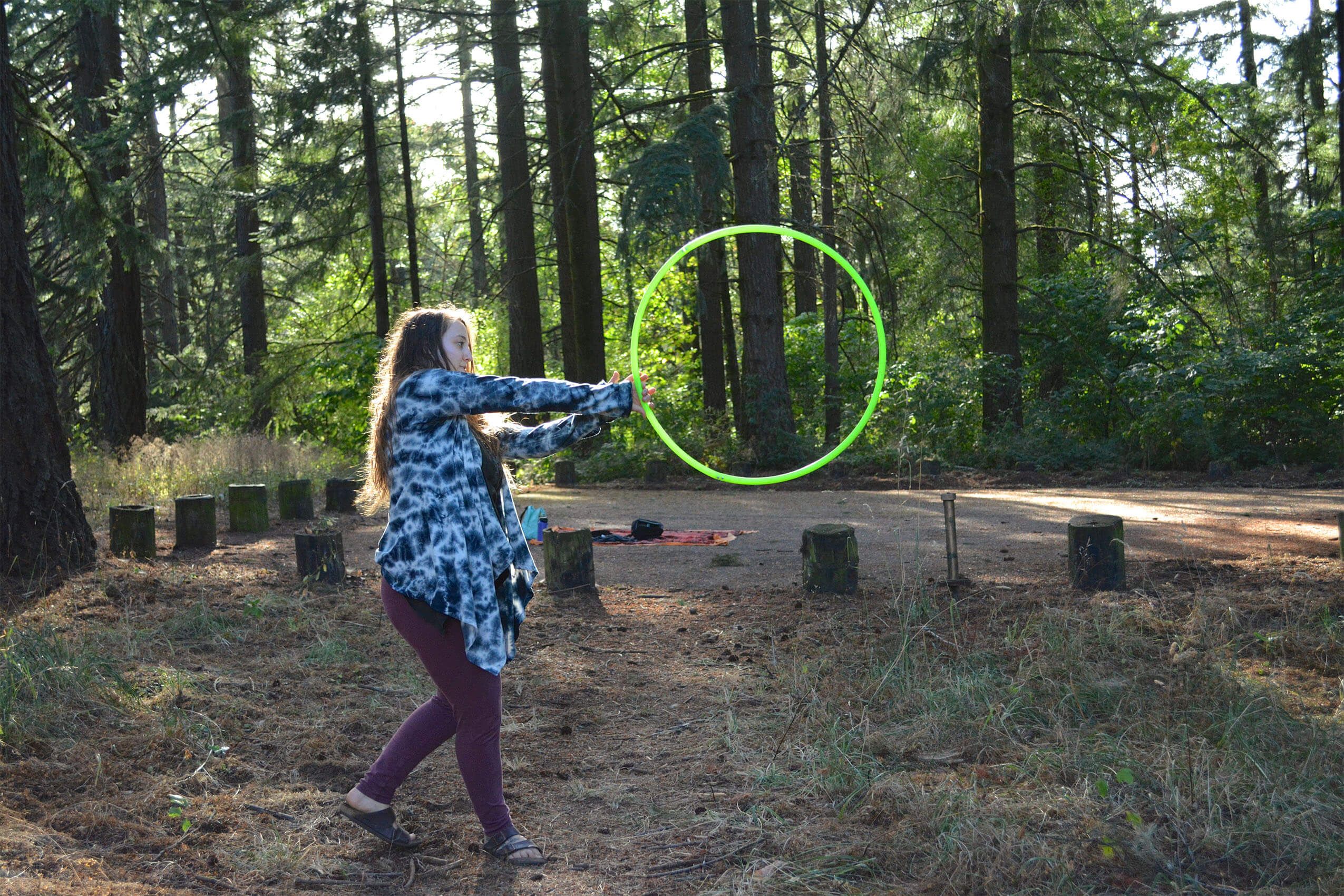 A person with a funky vibe stands in a forested area, holding a large green hoop on dry ground, surrounded by an earthy circle of wooden stumps. Soul Flower Blog: Hippie Style & Kind Living