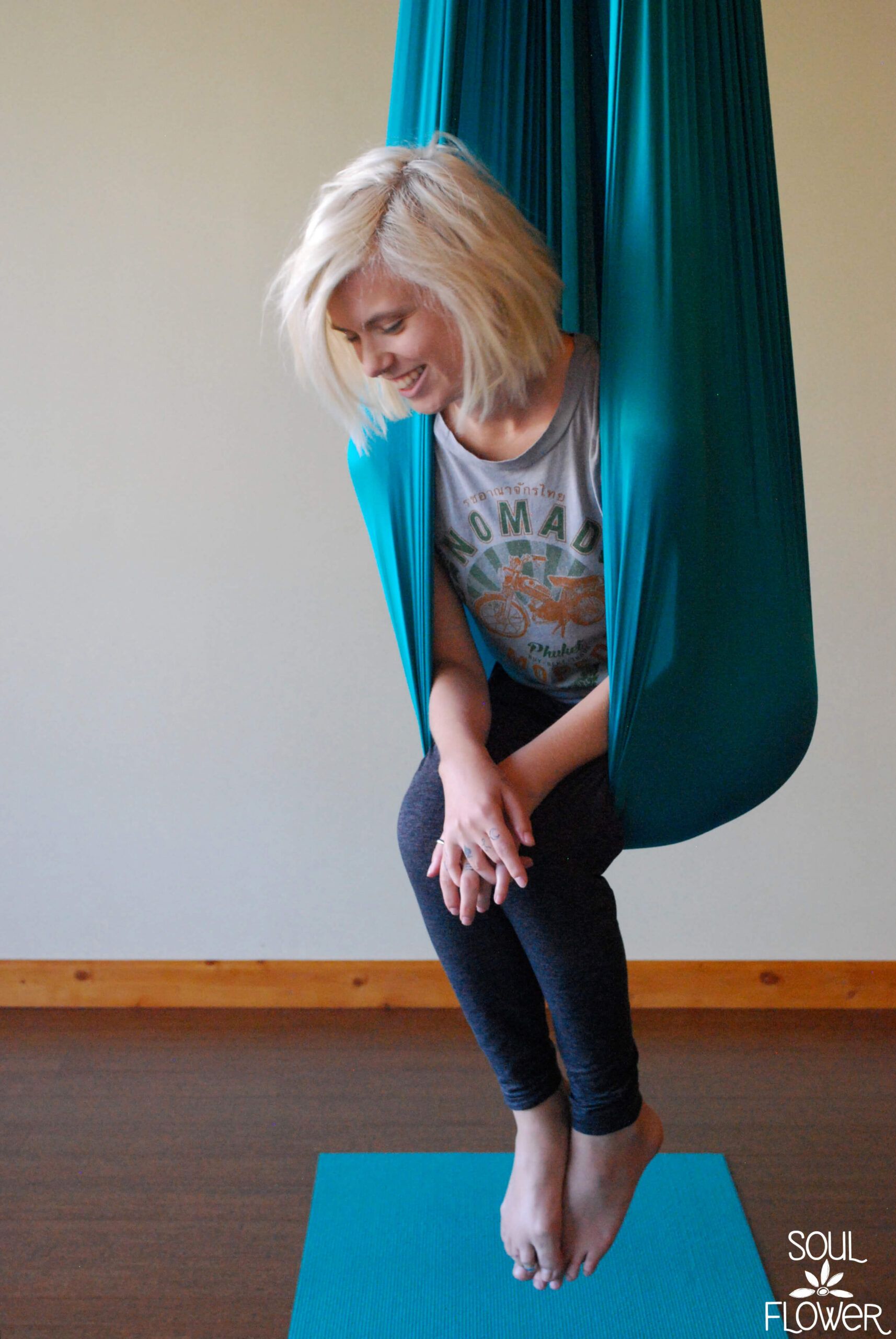 A person with blonde hair sits in an earthy teal aerial yoga hammock, smiling, with legs crossed, on a blue mat indoors. The text SOUL FLOWER appears in the bottom right corner. Soul Flower Blog: Hippie Style & Kind