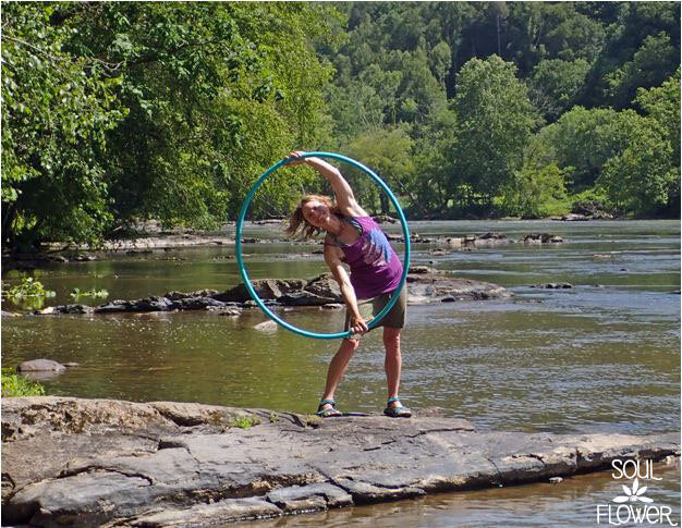 A person stands on a rocky, earthy riverbank holding a large blue hula hoop overhead, surrounded by trees and calm water. Hippie vibes abound. Soul Flower logo in the bottom right corner. Soul Flower Blog: Hippie Style &