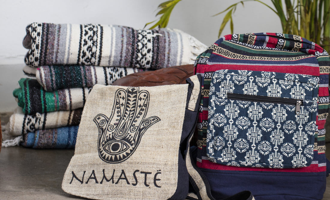 Stacked woven blankets, a small eco-friendly bag with a Namasté print and hamsa hand design, and a boho-patterned backpack are arranged on a concrete floor with plants in the background. Soul Flower Blog: Hippie Style & Kind Living