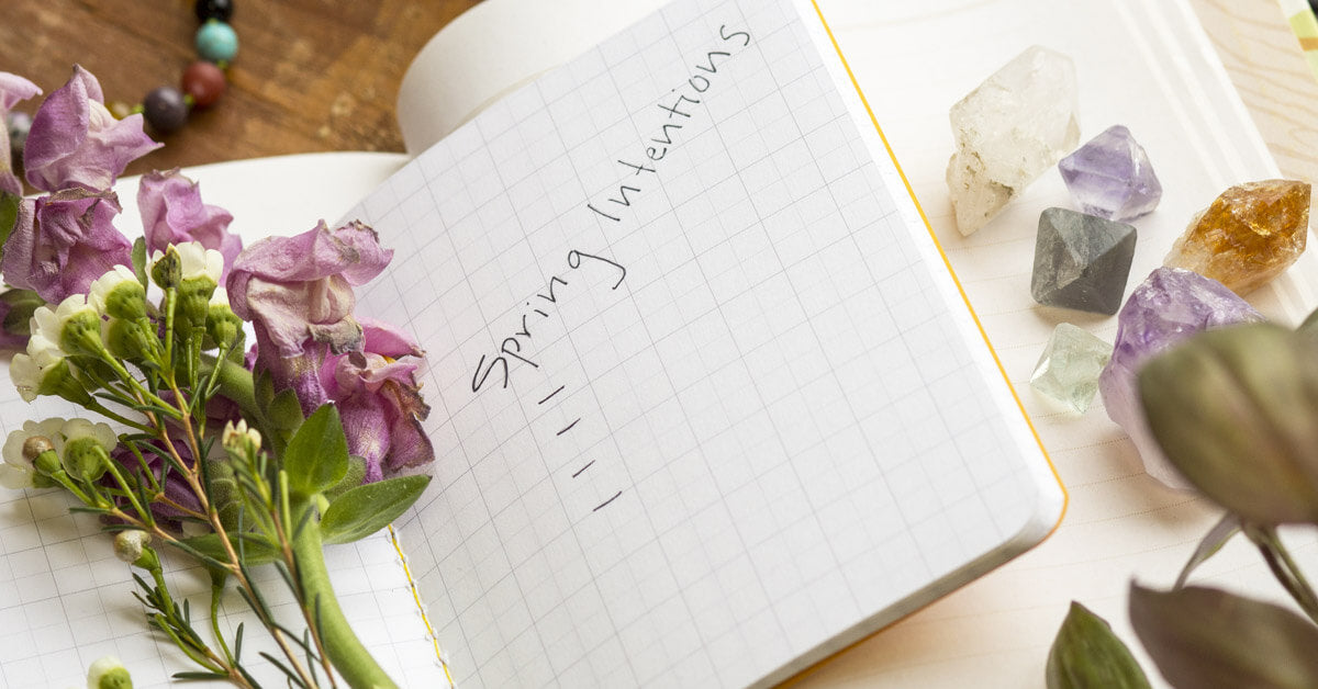 Open notebook with Spring Intentions written on the page, surrounded by purple flowers, green leaves, assorted crystals, and a beaded bracelet for a natural boho vibe on a wooden surface. Soul Flower Blog: Hippie Style & Kind Living
