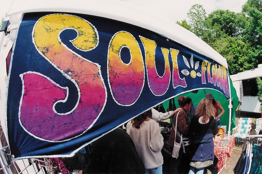 A market stall with a colorful tie-dye banner reading SOUL FLOWER welcomes shoppers for Small Business Saturday, with several people browsing unique items on tables under the tent. Soul Flower Blog: Hippie Style & Kind Living