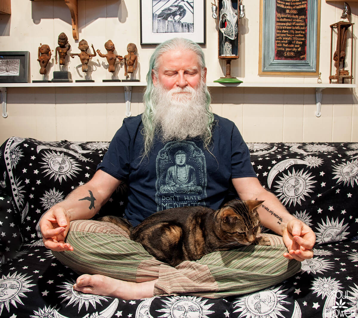 An older hippie man with long white hair and beard meditates cross-legged on a sofa, a cat in his lap, surrounded by earthy, eclectic art and figurines. Soul Flower Blog: Hippie Style & Kind Living