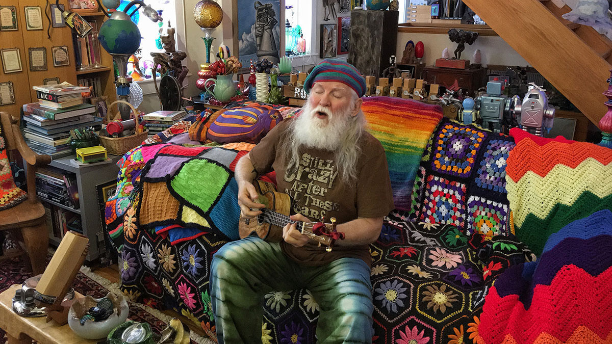 A dude with a long white beard sits on a colorful crocheted couch, playing a small stringed instrument in a room filled with eclectic, vibrant decor. Soul Flower Blog: Hippie Style & Kind Living