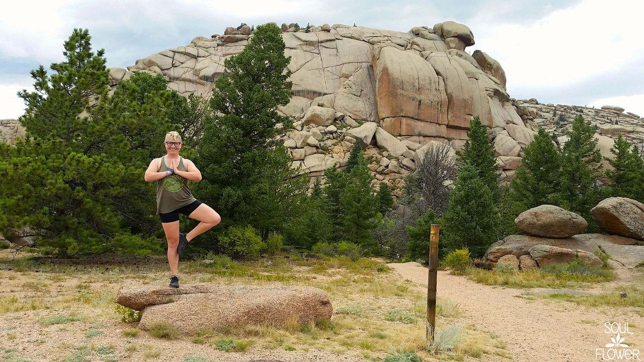 A person practices a yoga pose on a rock amidst pine trees and large boulders under a cloudy sky—Veda-what? Exploring Vedauwoo reveals peaceful moments like this in nature. Soul Flower Blog: Hippie Style & Kind Living