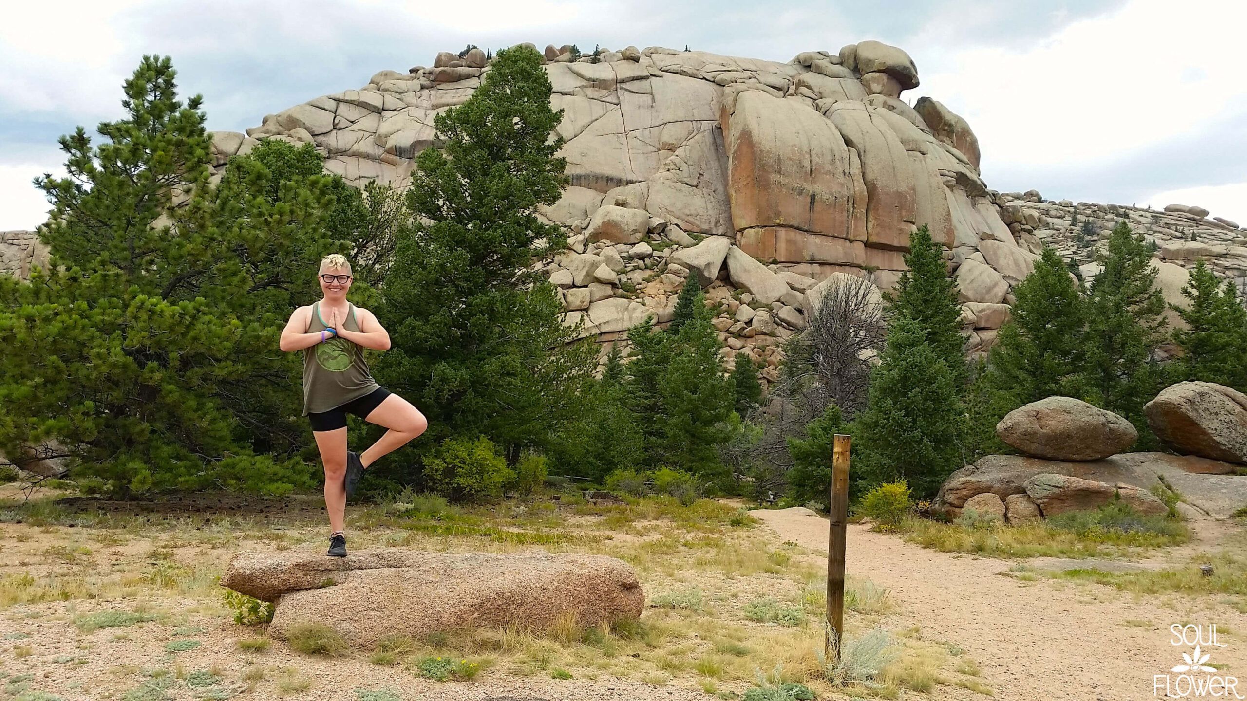 A person practices a yoga pose on a rock amidst pine trees and large boulders under a cloudy sky—Veda-what? Exploring Vedauwoo reveals peaceful moments like this in nature. Soul Flower Blog: Hippie Style & Kind Living