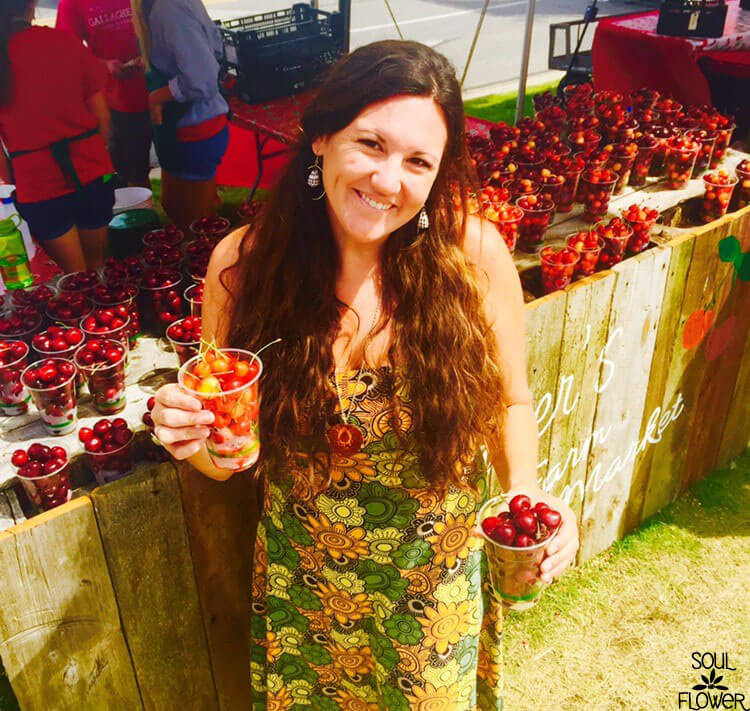 A woman in a boho floral dress holds a cup of cherries, standing by a wooden table filled with more cups of cherries at an earthy outdoor market. Soul Flower Blog: Hippie Style & Kind Living
