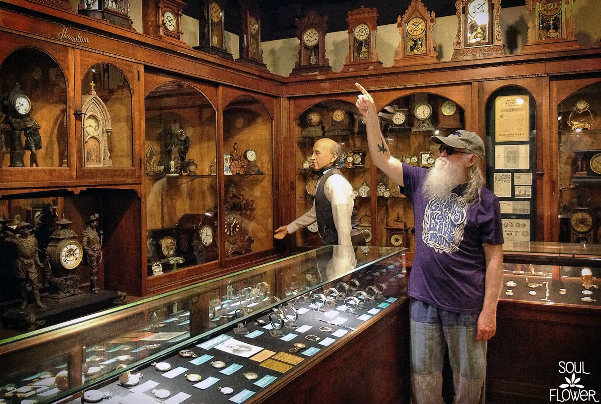 An older man with a long white beard points at a clock on a shelf in a museum room filled with antique clocks, as if he's On Becoming a Time Lord among the fascinating watches and display cases. Soul Flower Blog: Hippie