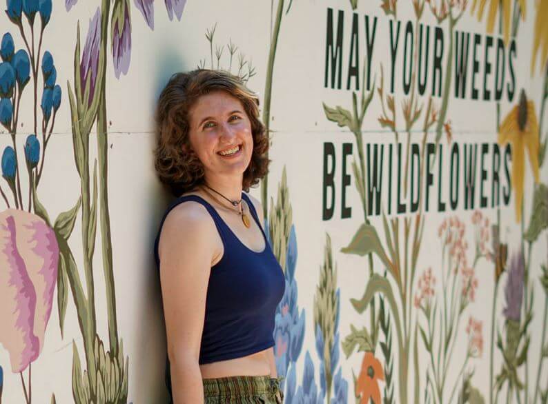 A person with short curly hair, wearing a navy crop top and green pants, leans against a boho mural that reads May your weeds be wildflowers with floral illustrations. Soul Flower Blog: Hippie Style & Kind Living