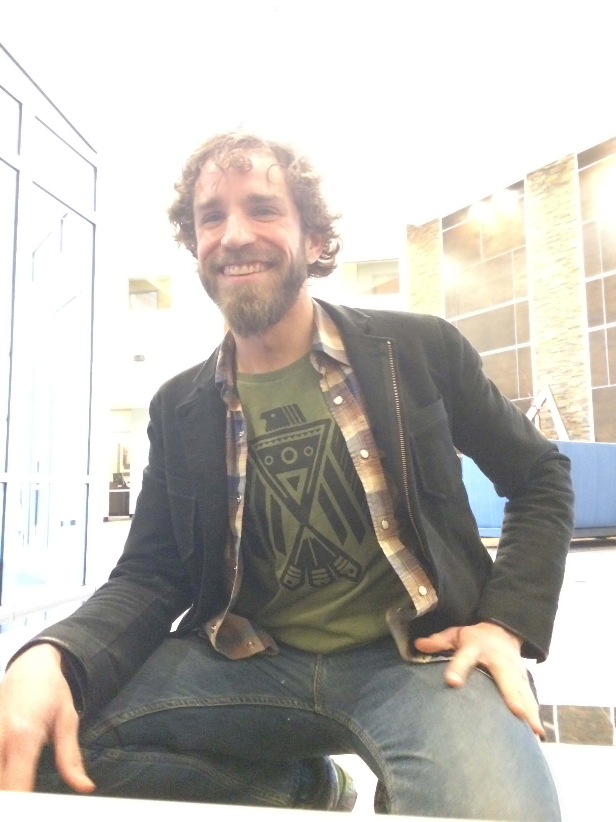 A man with curly hair and a beard sits indoors, smiling, wearing a black jacket, plaid shirt, and a green graphic T-shirt—his relaxed look hints at mindful living and a natural, boho-inspired style. Soul Flower Blog: Hippie Style & Kind Living