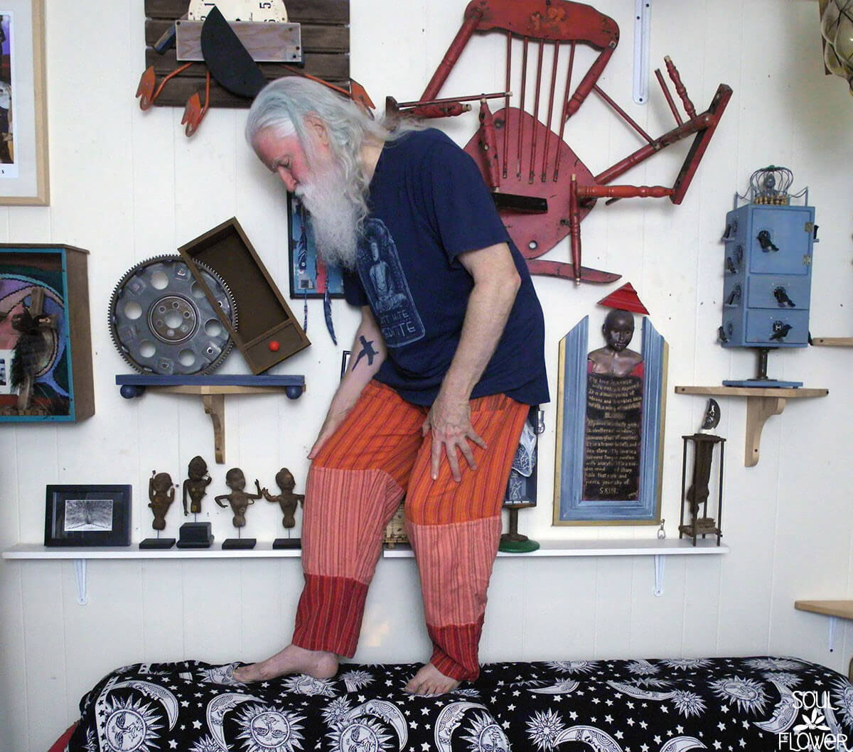 An older man with long white hair and beard stands barefoot on a patterned bed, surrounded by boho art, eclectic sculptures, and colorful wall decorations in a vibrant room. Soul Flower Blog: Hippie Style & Kind Living