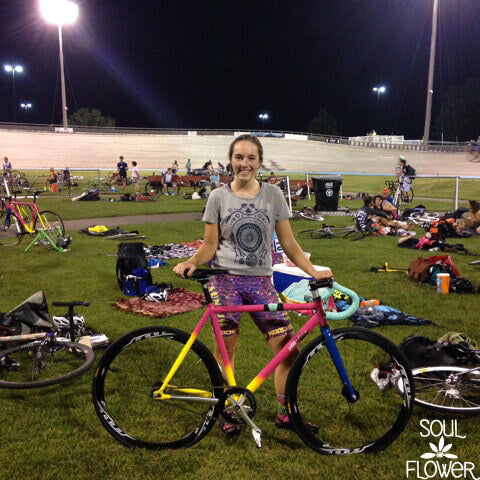 A person stands on a grassy field at a cycling track, holding a brightly colored bicycle—Finding Sangha in the Saddle. Soul Flower Blog: Hippie Style & Kind Living