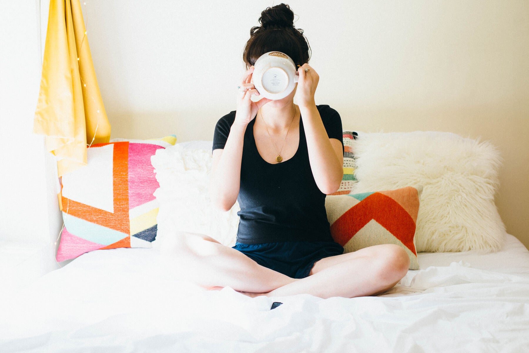Person sitting cross-legged on a bed, enjoying slow mornings while sipping from a large mug, with colorful pillows and a white fluffy cushion in the background. Soul Flower Blog: Hippie Style & Kind Living