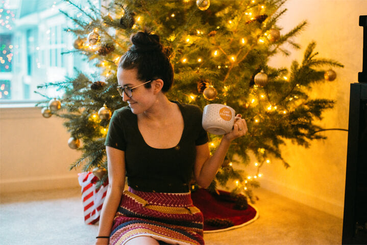 A woman with glasses sits on the floor in front of a decorated Christmas tree, holding a mug and smiling while looking to the side, exuding a cozy boho vibe. Soul Flower Blog: Hippie Style & Kind Living