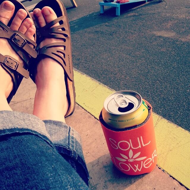 A person wearing brown sandals and jeans sits with crossed feet next to a can in an orange Soul Flower koozie on a sidewalk. Fan Photos! May-July 2015. Soul Flower Blog: Hippie Style & Kind Living