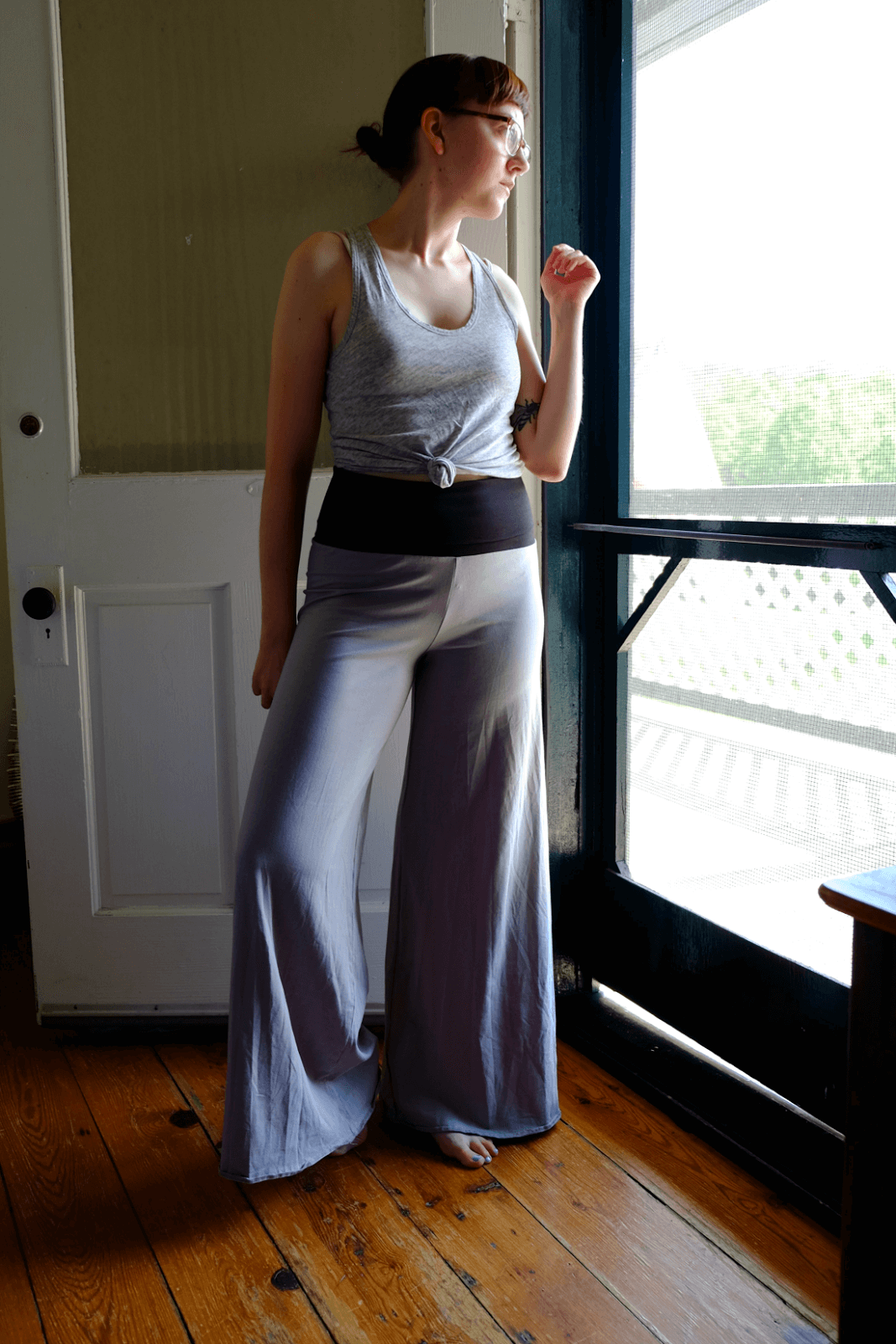 A kind woman in a sleeveless gray top and wide-legged boho pants stands barefoot by a door, looking out a window as natural light softly illuminates her. Soul Flower Blog: Hippie Style & Kind Living