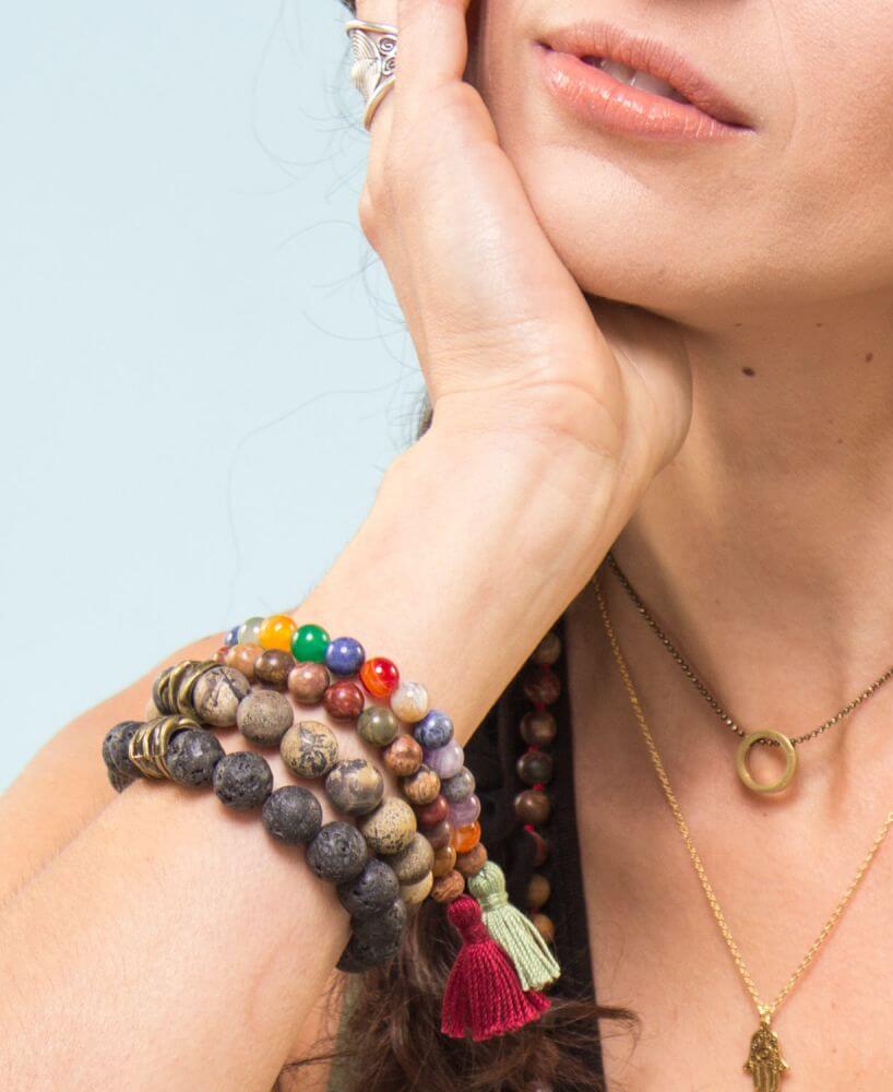 A woman with an earthy, boho vibe rests her chin on her hand, adorned with multiple beaded bracelets, necklaces, and a ring, set against a plain light blue background. Soul Flower Blog: Hippie Style & Kind Living