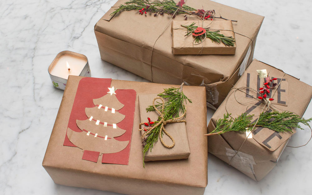 Four eco gifts wrapped in brown paper and decorated with greenery, twine, red berries, and a paper Christmas tree rest on a marble surface beside a lit candle. Soul Flower Blog: Hippie Style & Kind Living