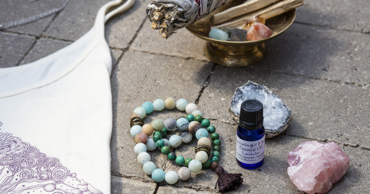 Assorted earthy items including beaded bracelets, a blue essential oil bottle, crystals, a bundle of sage, and a white cloth bag arranged on a stone surface for a boho touch. Soul Flower Blog: Hippie Style & Kind Living