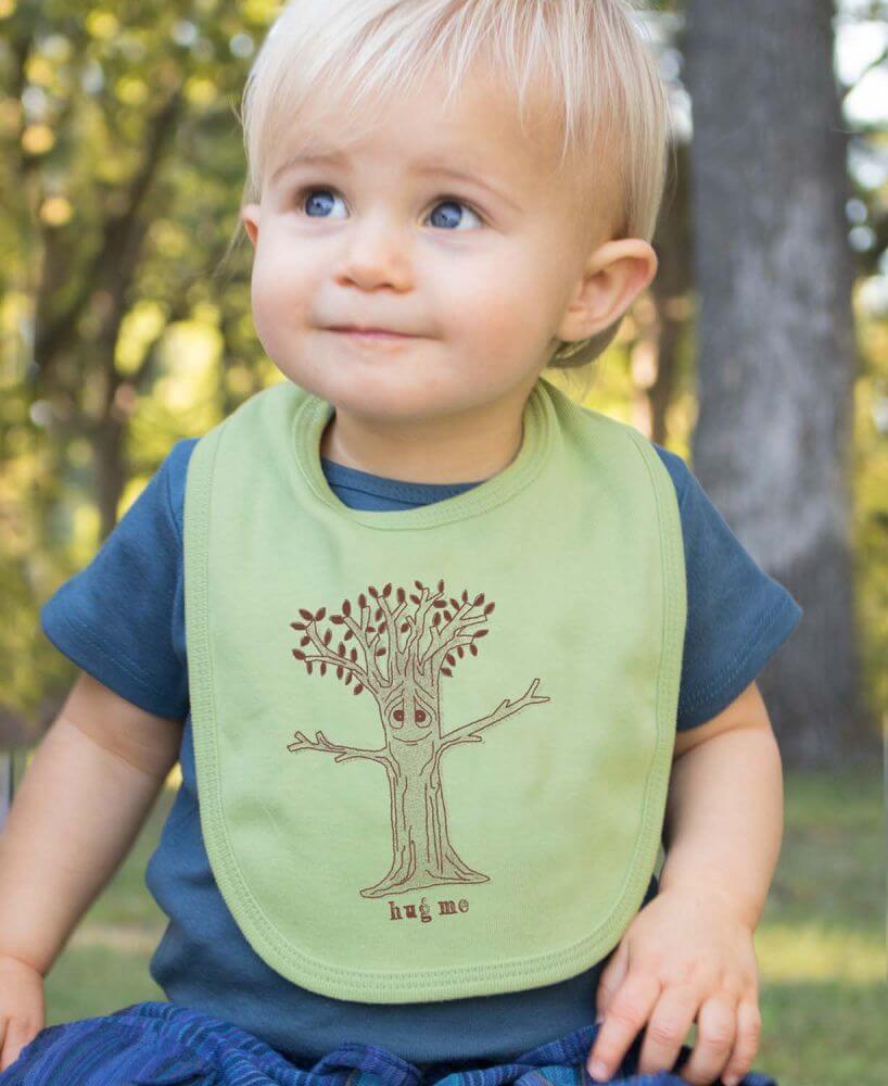 A toddler sits outdoors wearing a blue shirt and a green bib with a tree illustration and the words "hug me," making for adorable gifts for tree lovers. Soul Flower Blog: Hippie Style & Kind Living