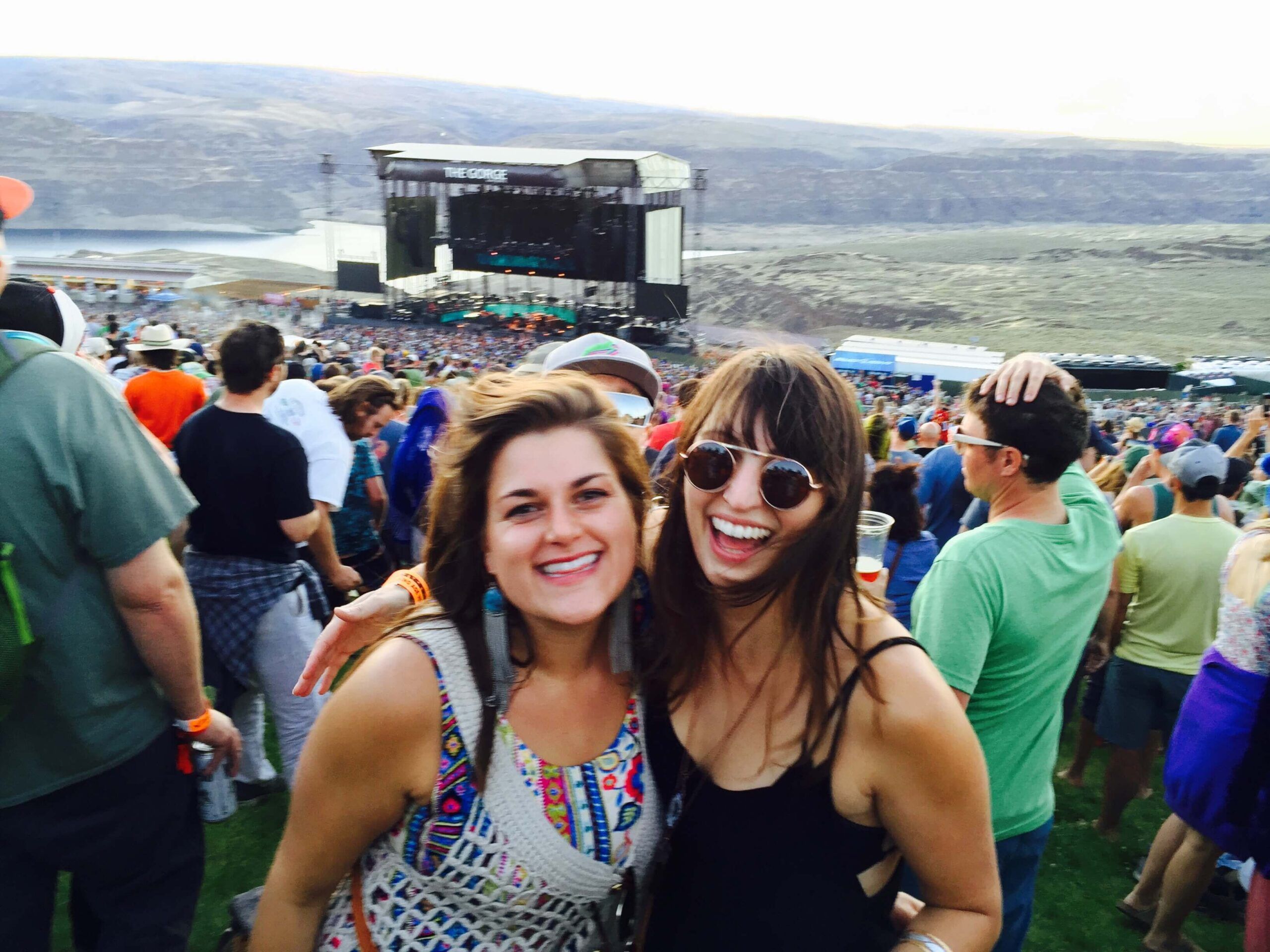 Two women smiling and posing for a photo at an outdoor concert, perfectly capturing what to wear to a music festival with the stage, crowd, and scenic landscape in the background. Soul Flower Blog: Hippie Style & Kind Living
