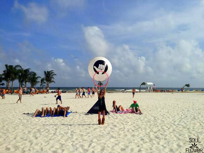 A woman stands on a sandy beach holding a pink hoop in front of a large, earthy circular art piece, with people sunbathing and palm trees in the background. Soul Flower Blog: Hippie Style & Kind Living