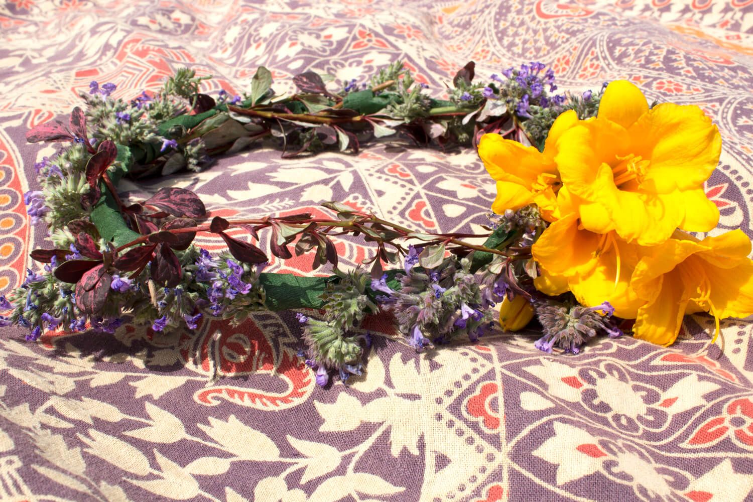 A Hippie Flower Crown made of yellow flowers, purple blossoms, and greenery lies on a patterned fabric background. Soul Flower Blog: Hippie Style & Kind Living