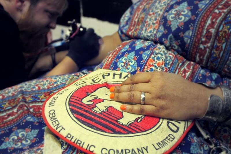 A person with orange-painted nails rests their hand on a boho fabric patch while someone tattoos their side in the background. Soul Flower Blog: Hippie Style & Kind Living