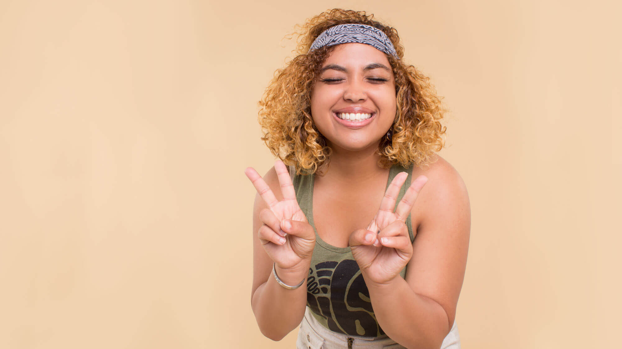 Smiling, kind woman with curly hair and a funky headband making two peace signs with both hands in front of a plain beige background. Soul Flower Blog: Hippie Style & Kind Living