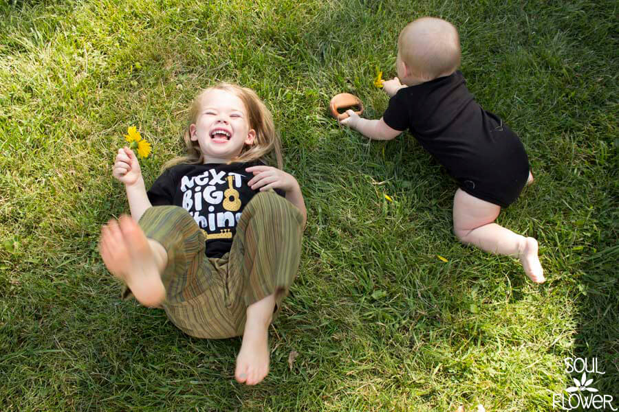 Meet Our Naturally Cute Little Buds: Two young children play on green grass; one laughs while holding a yellow flower, the other crawls nearby. Both are dressed in casual clothes and brimming with joy. Soul Flower Blog: