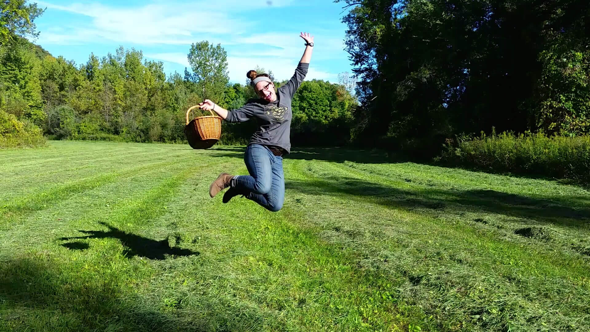 A person with a bun hairstyle jumps in the air on a grassy field, holding a wicker basket; trees and blue sky set the scene, echoing the spirit of "You Belong Among the Wild Fox Grapes. Soul Flower Blog: Hippie Style & Kind Living