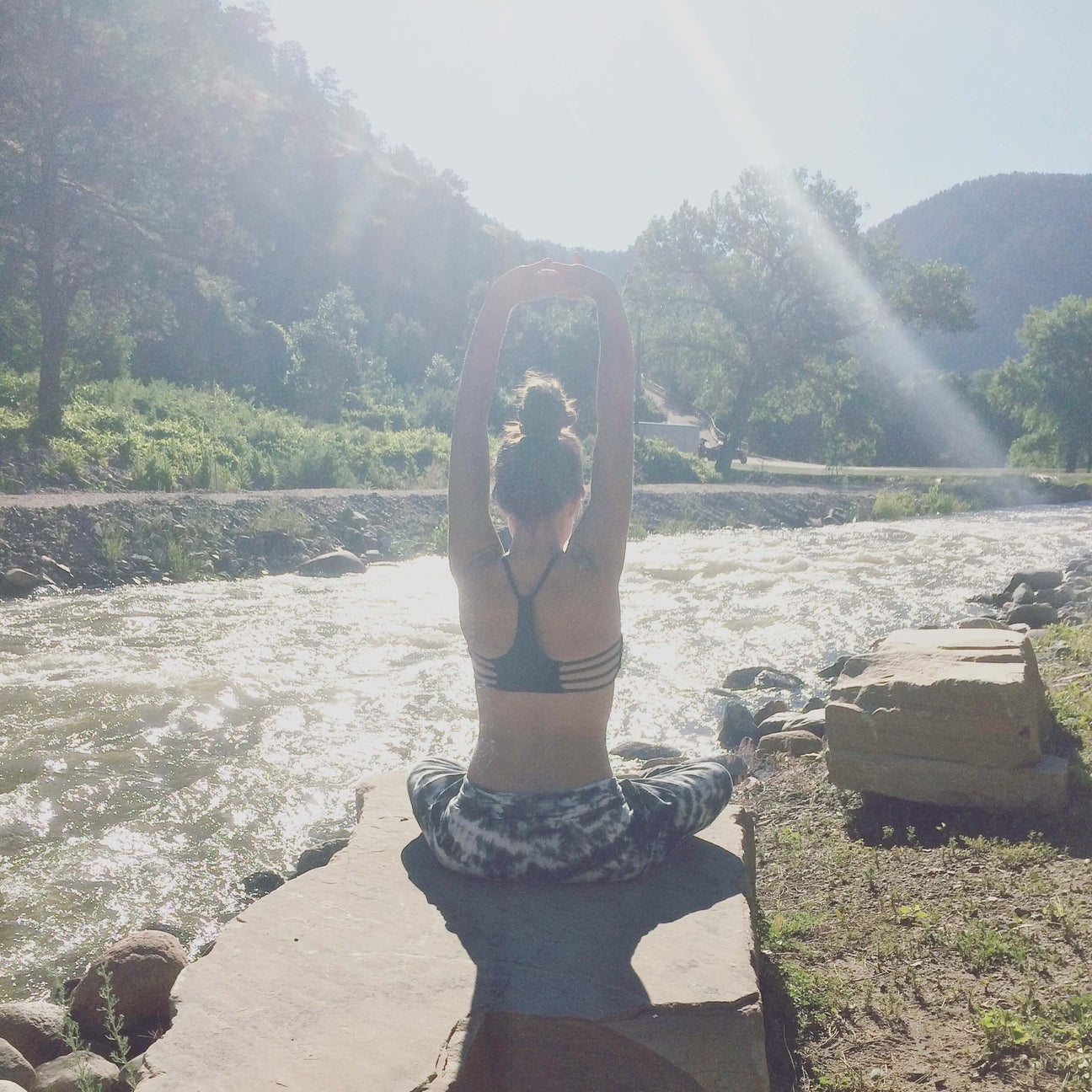 Person sitting cross-legged on a rock by a river, stretching arms overhead, with sunlight streaming through trees—capturing the calm you’ll feel by following these 7 Tips for Having a Stress-Free Wedding Day. Soul Flower Blog: Hippie Style & Kind Living