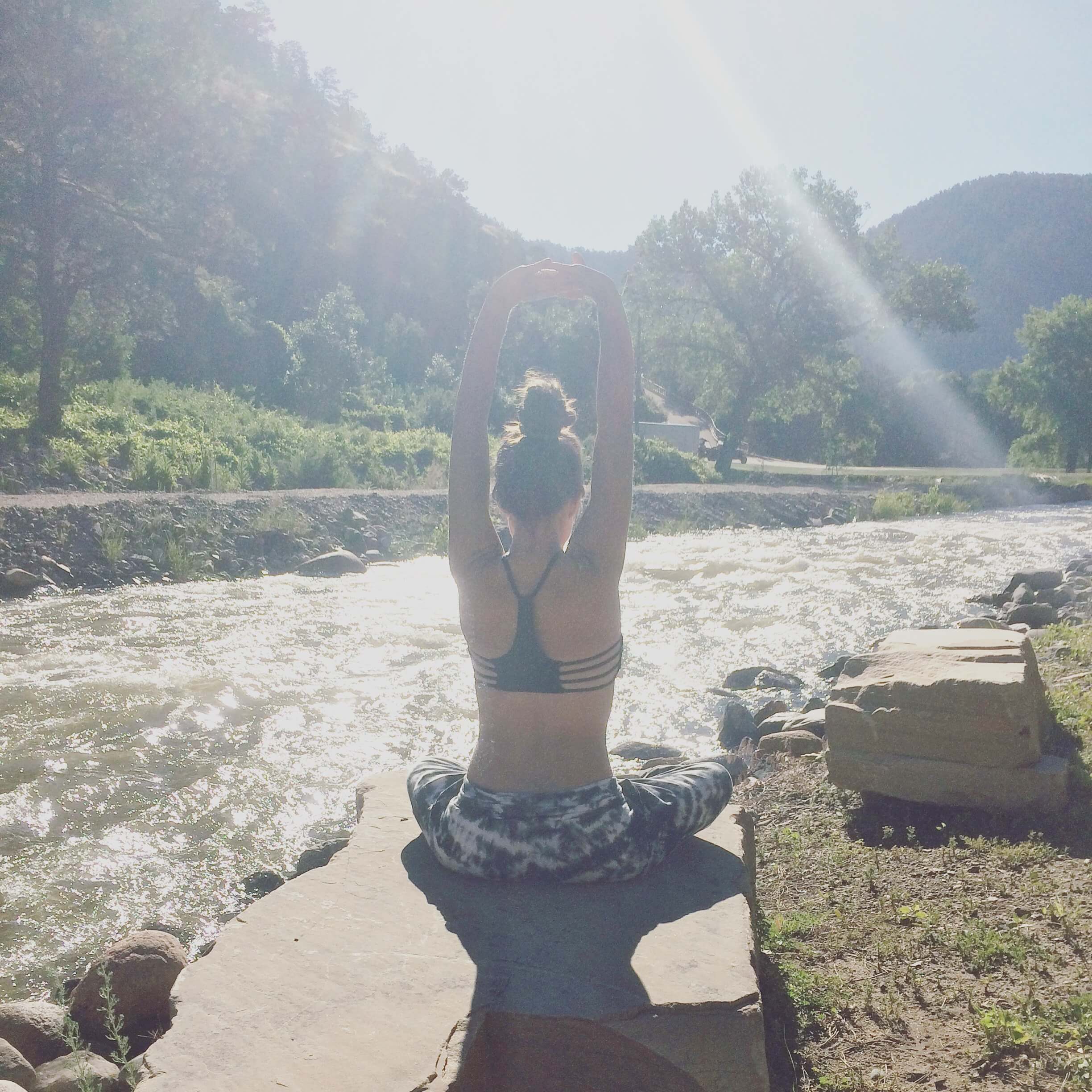 Person sitting cross-legged on a rock by a river, stretching arms overhead, with sunlight streaming through trees—capturing the calm you’ll feel by following these 7 Tips for Having a Stress-Free Wedding Day. Soul Flower Blog: Hippie Style & Kind Living