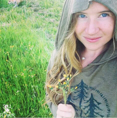 A woman in a hooded green sweatshirt with a tree design stands in a grassy field, holding small yellow flowers and smiling at the camera for an earthy, boho vibe. Soul Flower Blog: Hippie Style & Kind Living