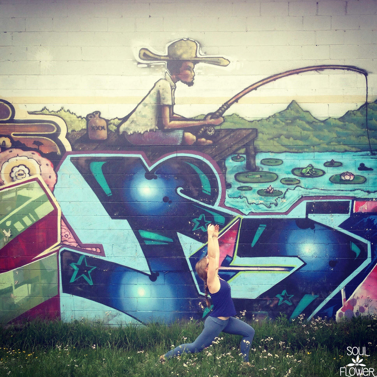 A person does a yoga pose in front of a colorful, earthy mural depicting a person fishing by a lake, with boho graffiti and landscape elements. Soul Flower Blog: Hippie Style & Kind Living